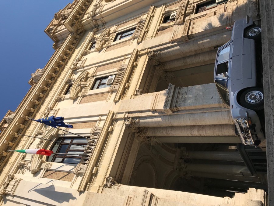 Low angle shot of a vintage grey car beside a neoclassical Roman building with Italian and EU flags